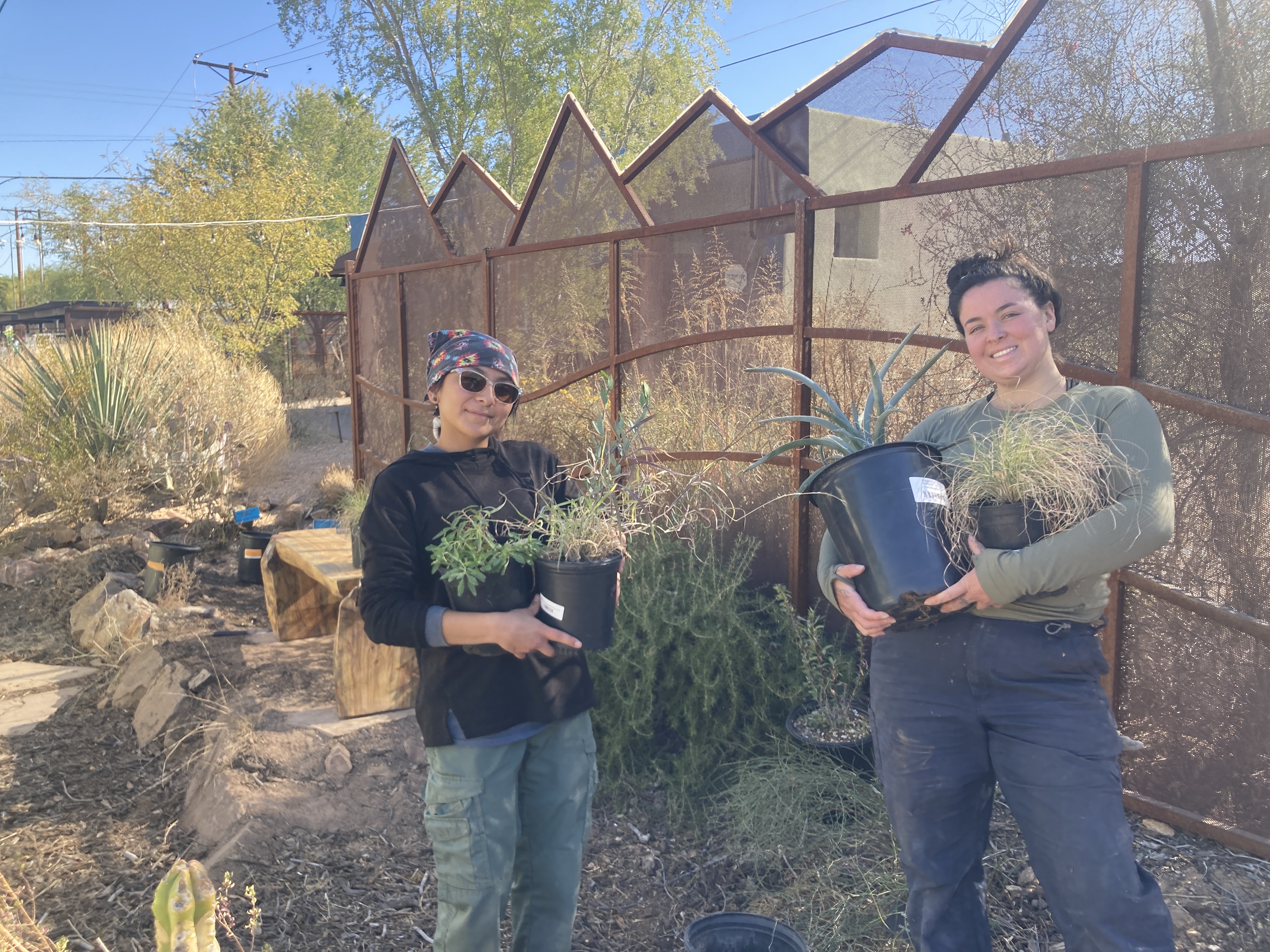 Two young women hold pots of plants in front of a garden