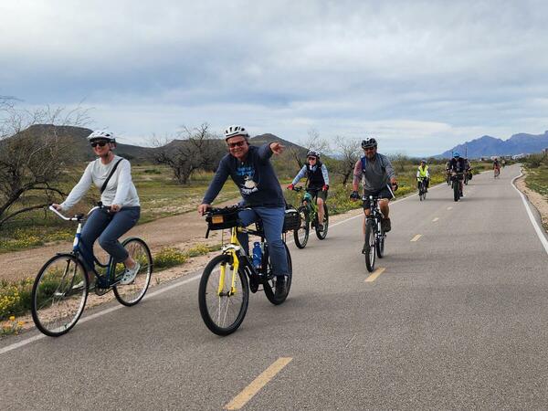 A group of people ride bikes on a bike path -- the one in front is smiling and pointing at the camera