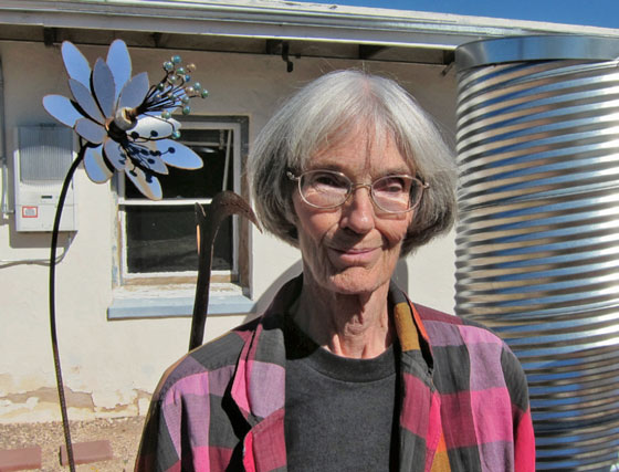 A light skinned older woman with a grey bob stands in front of an adobe house, a metal cistern, and a metal flower sculpture