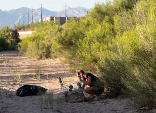 A person squats in a dry river bed, digging up an upside down shopping cart