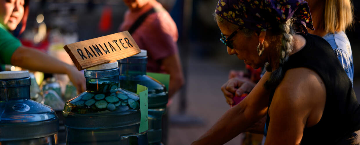 A person in a tank top and purple head scarf dispenses water from a jug labeled rainwater