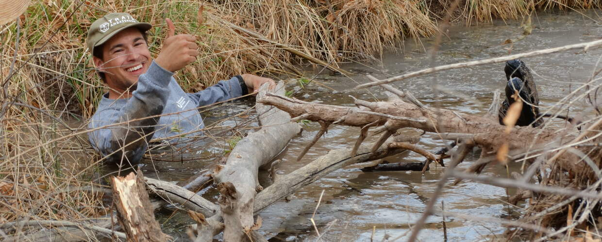 A man sits in a creek covered in logs, laughing and giving a thumbs up