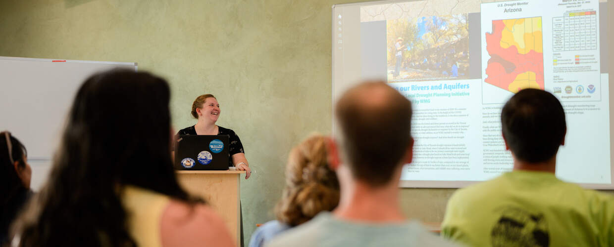 Teacher smiles in front of a class of adults facing a projection screen
