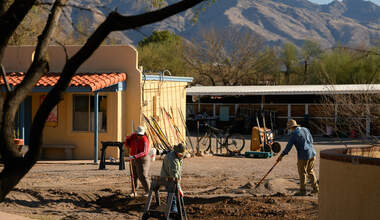 a group of people use shovels and rakes to shape the ground for basins in front of a yellow mission style building and a distant mountain range
