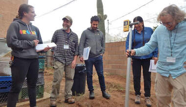 A group of five people smile and stand around an in construction earthwork while holding notebooks. One of them is holding a shovel